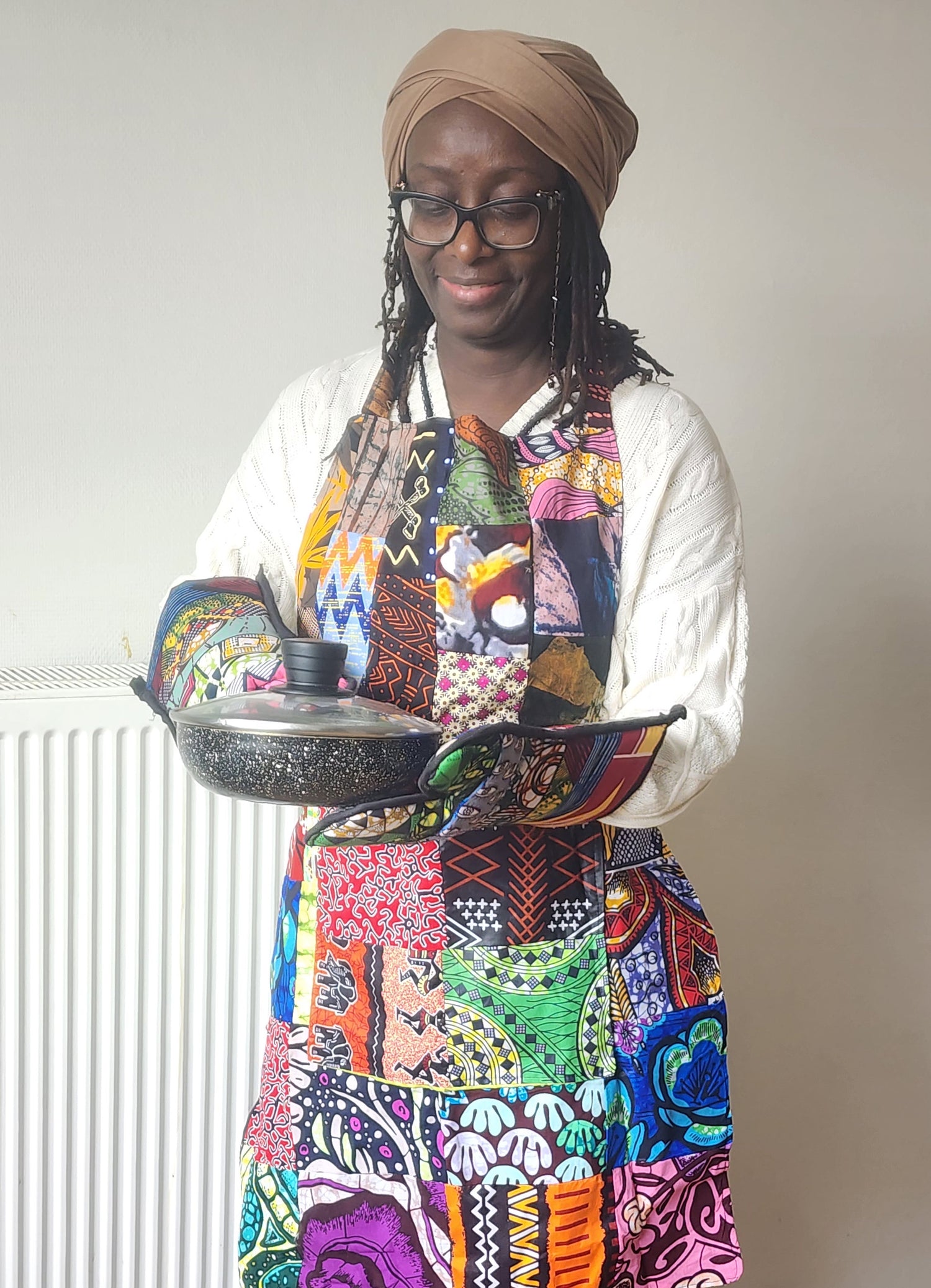 Woman wearing a colorful apron and glasses, holding a pot against a plain background