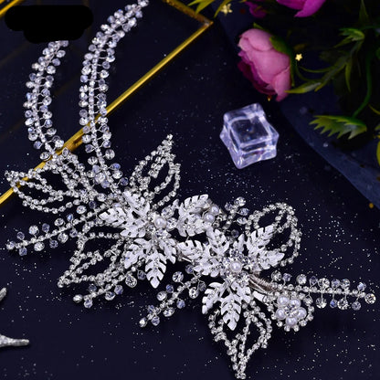 Silver crystal vine bridal hairpiece with leaf and pearl detailing displayed flat on black background with flowers.