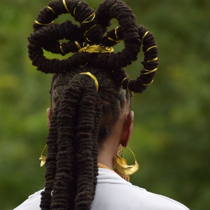 Person with braided hair styled in a decorative manner with gold accents, wearing gold earrings against a blurred green background.