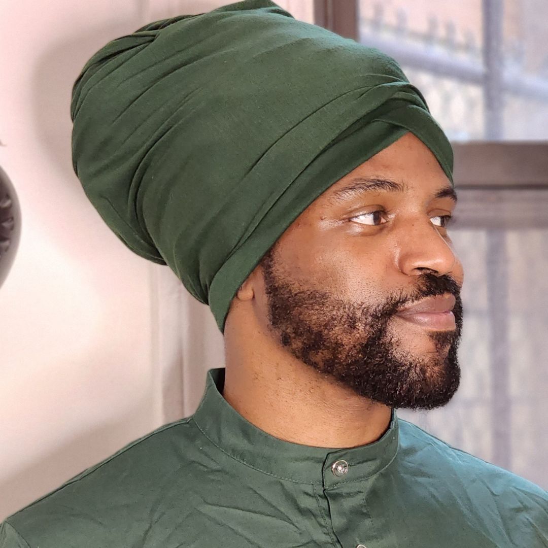 A man wearing a dark green turban and a matching green button-up shirt, shown in a close-up profile view against an indoor background.