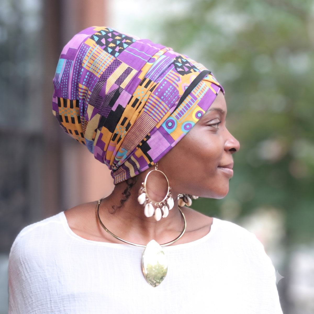A woman in profile wearing a vibrant purple, yellow, and black geometric patterned headwrap, styled with gold statement jewelry and a white top against a softly blurred outdoor background.