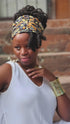 Woman wearing the Regal Adornment Set, including a gold-tone cuff bracelet, cowrie shell earrings, and a patterned headwrap. A stylish accessory for locs and braids.